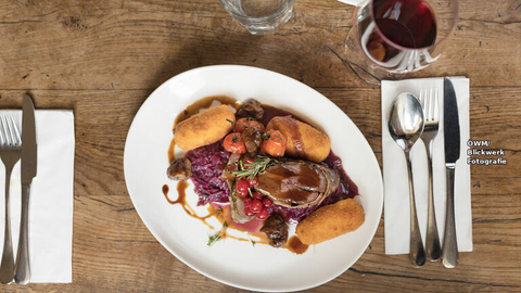 Steirisches Wildgericht mit Rotkraut, Kroketten und einem Glas Rotwein auf rustikalem Holztisch – Weinempfehlungen zu Wildgerichten von Weinhandel Steiermarkt.